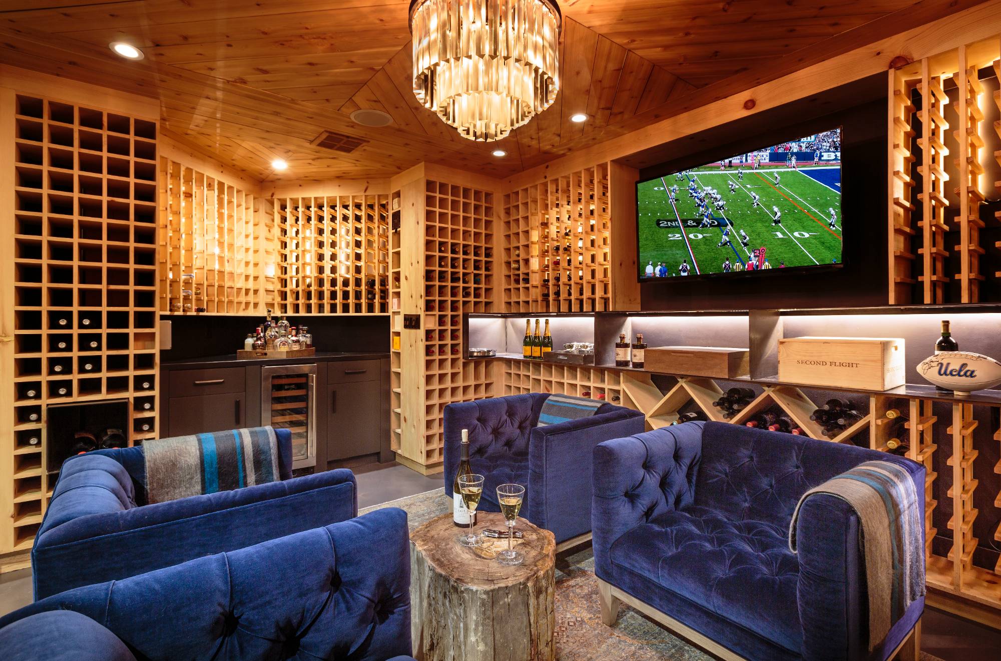 Custom climate-controlled wine basement built by ESRL Development, floor-to-ceiling cedar wine racking holding hundreds of bottles, built-in refrigeration unit, mounted flat-screen television, crystal chandelier, and tufted blue velvet lounge seating. Brentwood Layton Drive residence.