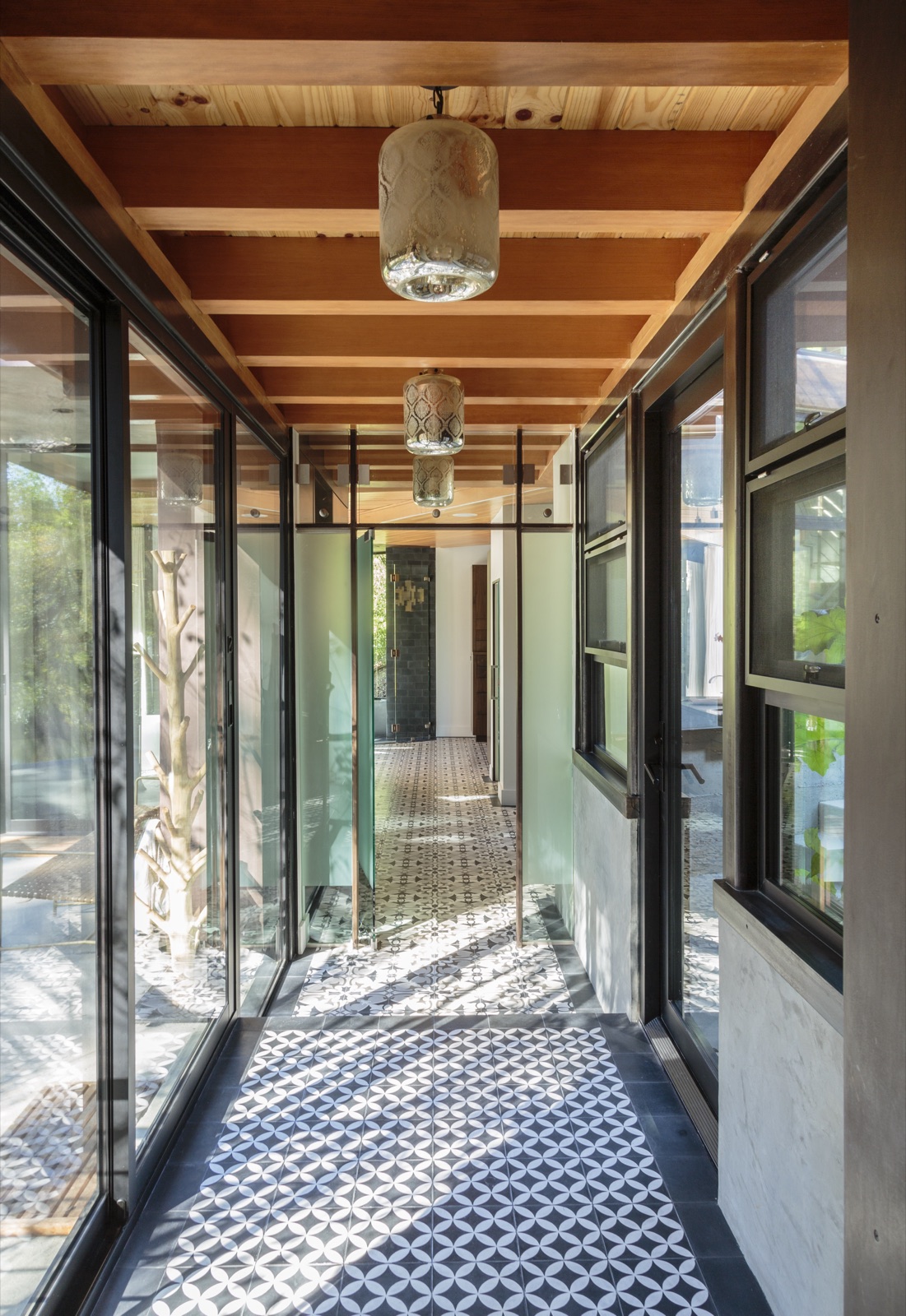 Glass-walled gallery hallway connecting the addition to the original house, patterned cement-tile floor, exposed cedar beam ceiling, blackened-steel windows on both sides. Brentwood Layton Drive residence, ESRL Development.