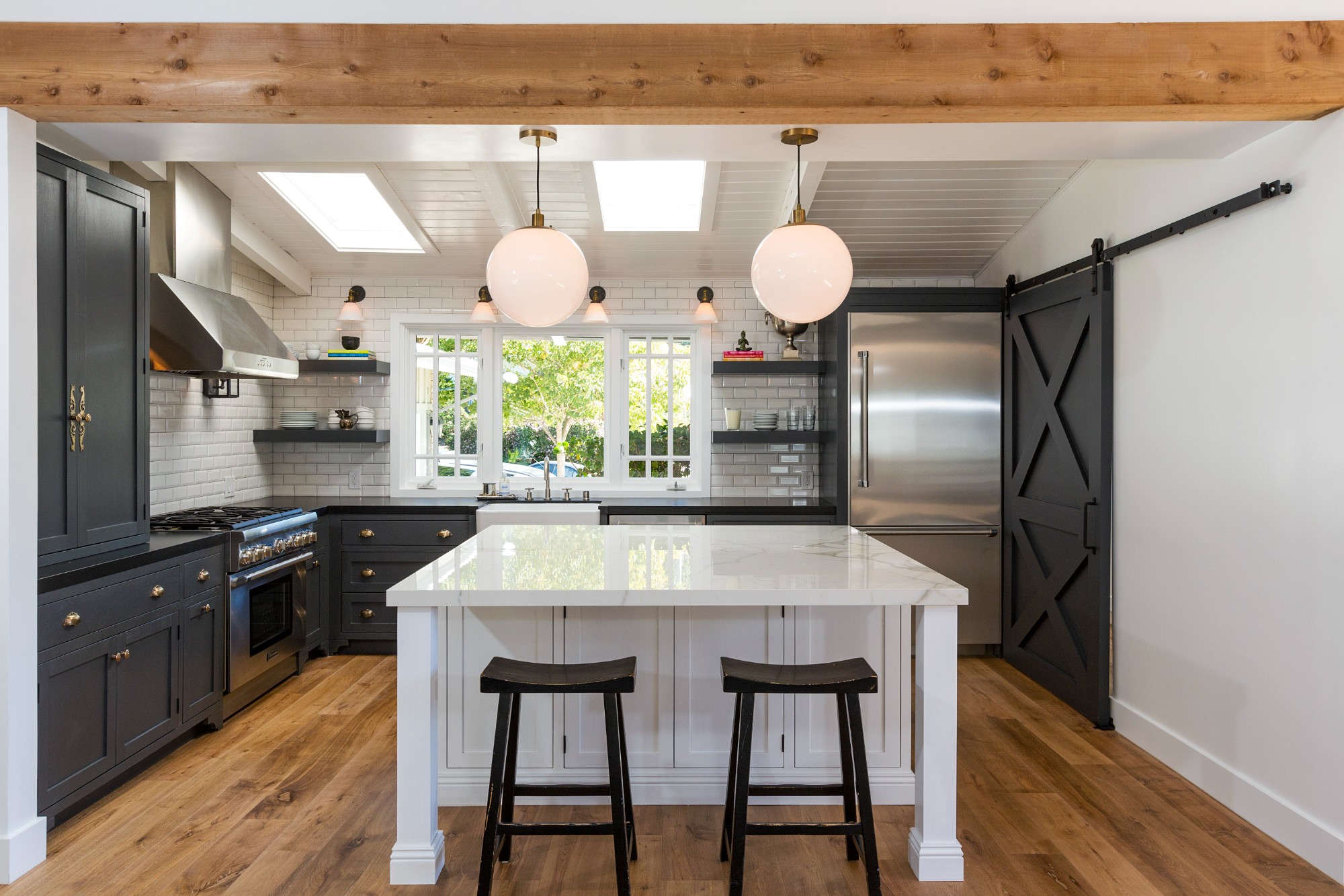 Pacific Palisades beach house kitchen remodel by ESRL Development, charcoal shaker cabinetry with brass cup pulls, Calacatta marble waterfall island, white subway tile backsplash, sliding barn door, skylit vaulted ceiling, exposed wood beam.