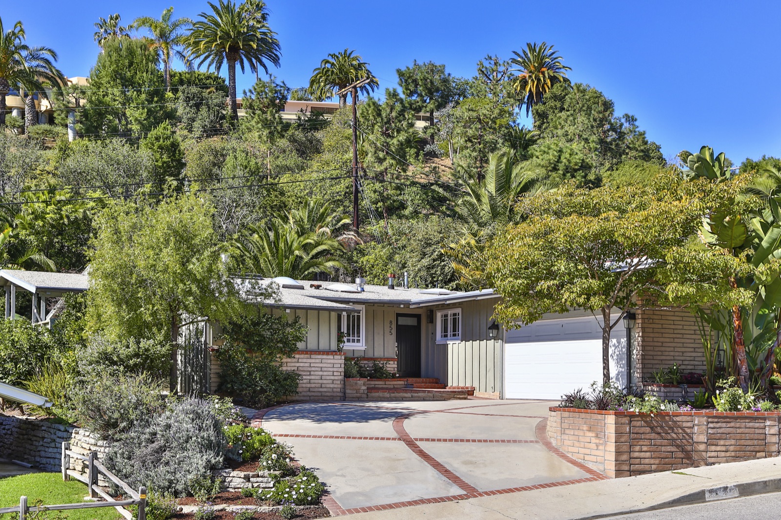 855 Jacon Way Pacific Palisades beach house by ESRL Development, front elevation of the single-story California cottage with restored board-and-batten siding, brick-edged concrete driveway, two-car garage, mature palms and the Palisades hillside rising behind.