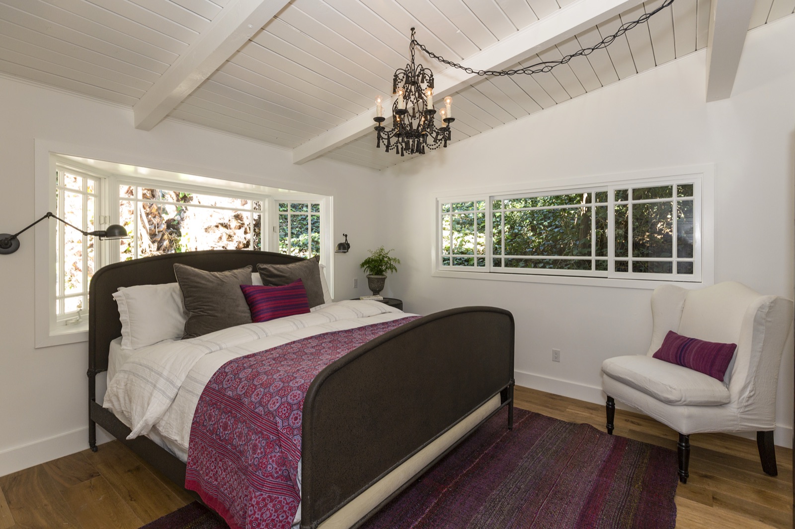 Master bedroom at 855 Jacon Way Pacific Palisades, exposed beam ceiling, wrought-iron chandelier, custom dark-upholstered four-poster bed, layered linens, and a wraparound bay of casement windows opening to the garden. ESRL Development beach house remodel.