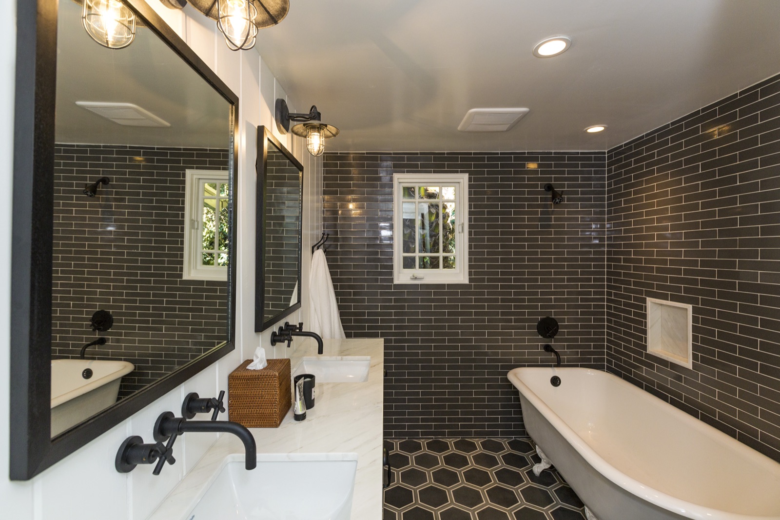 Master bathroom at 855 Jacon Way Pacific Palisades, cast-iron clawfoot soaking tub under a steel-cage industrial sconce, black subway-tile walls with white grout, hex-tile floor, marble vanity, framed black mirrors. Coastal master bath remodel by ESRL Development.