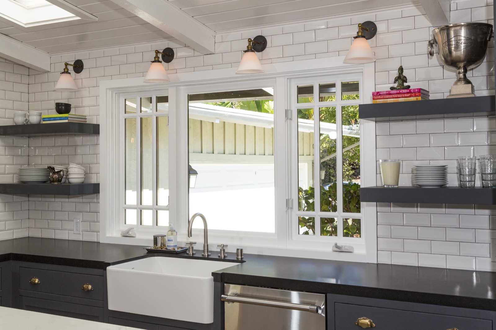 Kitchen detail at 855 Jacon Way Pacific Palisades, fireclay apron-front farmhouse sink, polished-nickel bridge faucet, white subway-tile walls with dark grout, custom open dark shelving styled with stoneware, four matching schoolhouse wall sconces overhead. ESRL Development.