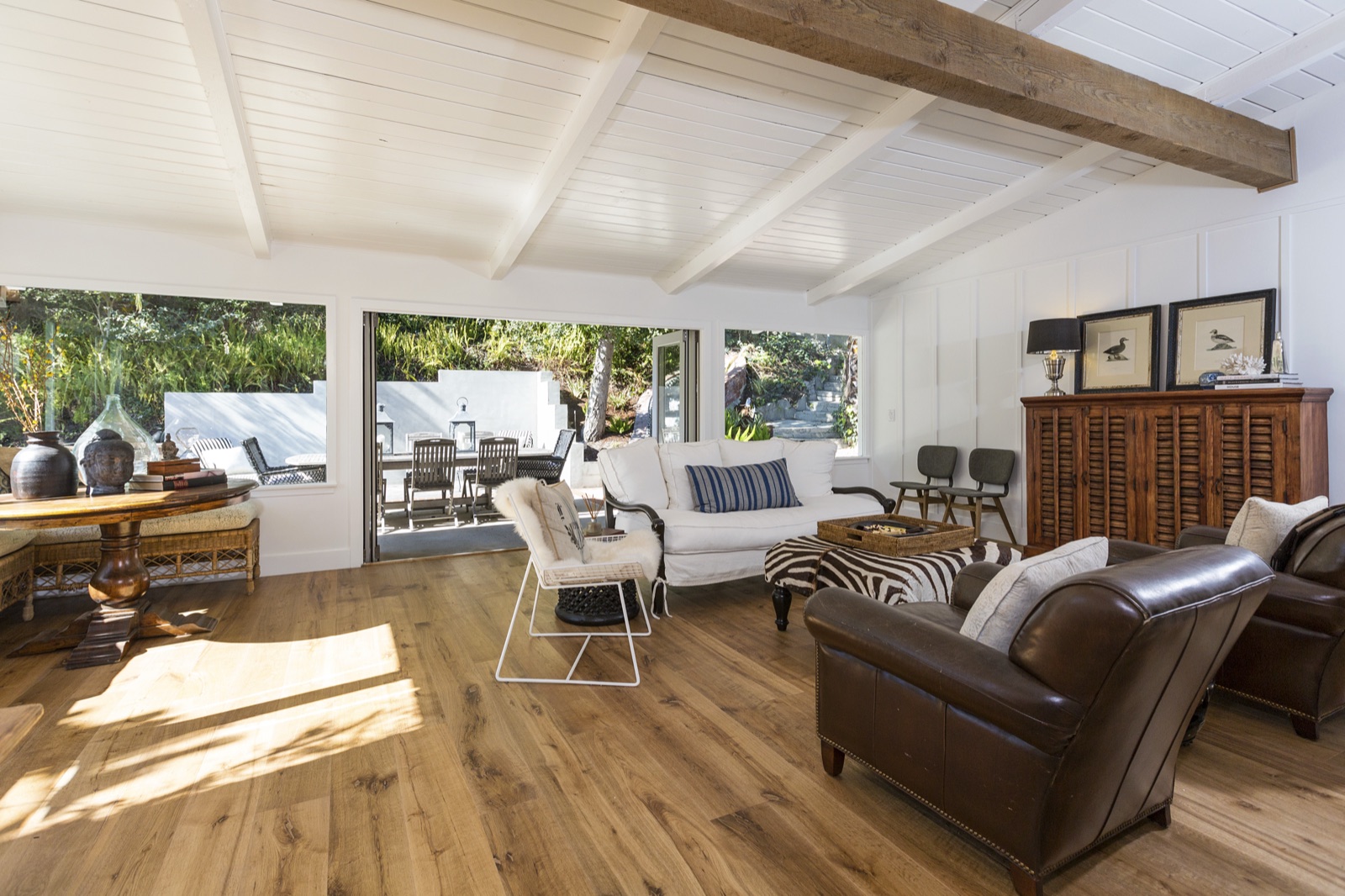 Great room at 855 Jacon Way Pacific Palisades opening to the backyard through full-height sliders, wide-plank white oak floors, white-painted beamed and shiplap ceiling, dining table beneath, deep leather club chair, indoor-outdoor flow. ESRL Development.