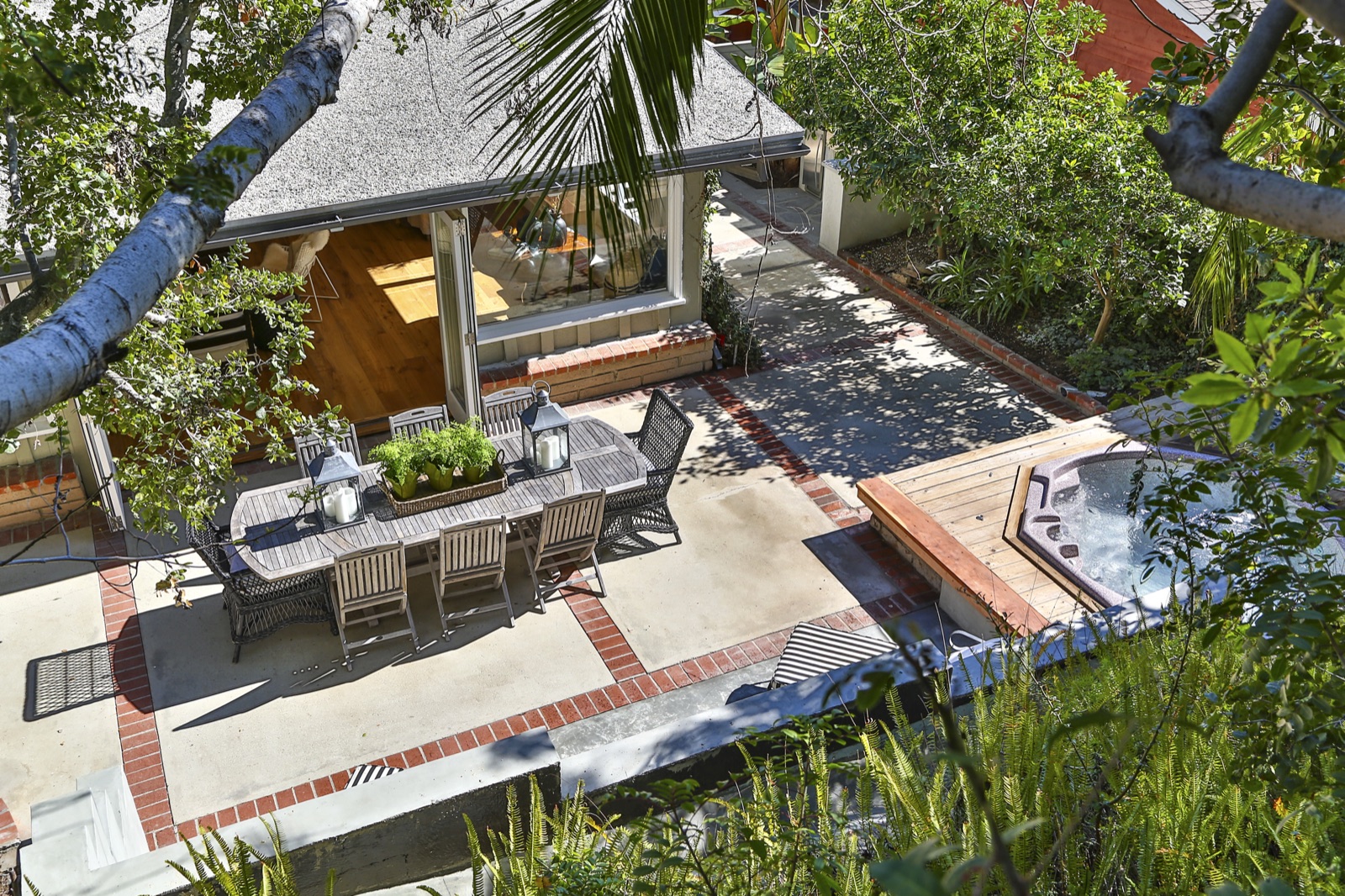 Aerial view of the backyard at 855 Jacon Way Pacific Palisades, outdoor dining cabana with shingled roof, hand-laid brick patio with banded inlay around the dining table, sunken hot-tub spa beside it, tucked into canyon greenery on the Palisades hillside. ESRL Development beach house remodel.